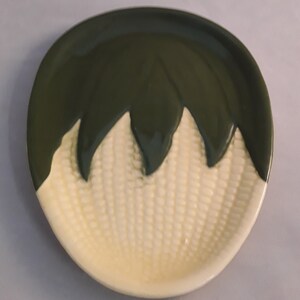 May include: A ceramic dish shaped like an ear of corn. The dish is white with green details and has a textured surface.