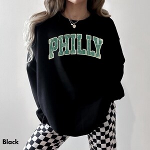 Op de afbeelding: Zwart sweatshirt met het woord "PHILLY" in verweerde groene letters. Het sweatshirt wordt gecombineerd met een zwart-wit geruite broek. Het woord "Black" staat linksonder in de afbeelding.