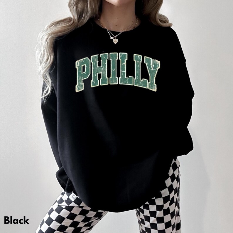 Op de afbeelding: Zwart sweatshirt met het woord "PHILLY" in verweerde groene letters. Het sweatshirt wordt gecombineerd met een zwart-wit geruite broek. Het woord "Black" staat linksonder in de afbeelding.