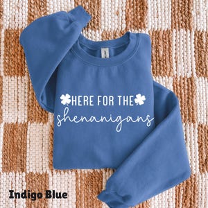 May include: Indigo blue sweatshirt with the text "Here for the shenanigans" in white script and shamrock accents. The sweatshirt is folded on a textured, neutral-toned surface.