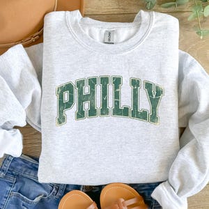 Op de afbeelding: Een lichtgrijs sweatshirt met het woord "PHILLY" in verweerde groene letters. Het sweatshirt is opgevouwen, met de mouwen zichtbaar. Het woord "Ash" staat linksonder afgedrukt. Het wordt gecombineerd met een spijkerbroek en sandalen.