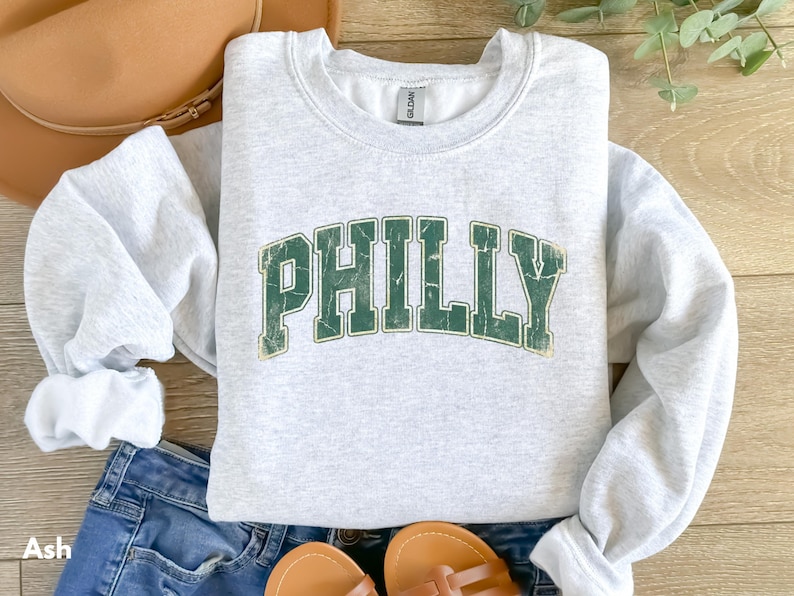 Op de afbeelding: Een lichtgrijs sweatshirt met het woord "PHILLY" in verweerde groene letters. Het sweatshirt is opgevouwen, met de mouwen zichtbaar. Het woord "Ash" staat linksonder afgedrukt. Het wordt gecombineerd met een spijkerbroek en sandalen.