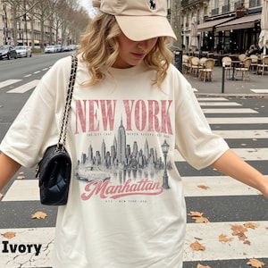 May include: An ivory t-shirt featuring a graphic print of the New York City skyline, a car, and a street lamp. The text reads NEW YORK, THE CITY THAT NEVER SLEEPS EST 1624, Manhattan, and NYC NEW YORK USA. A black quilted shoulder bag is worn on the side.