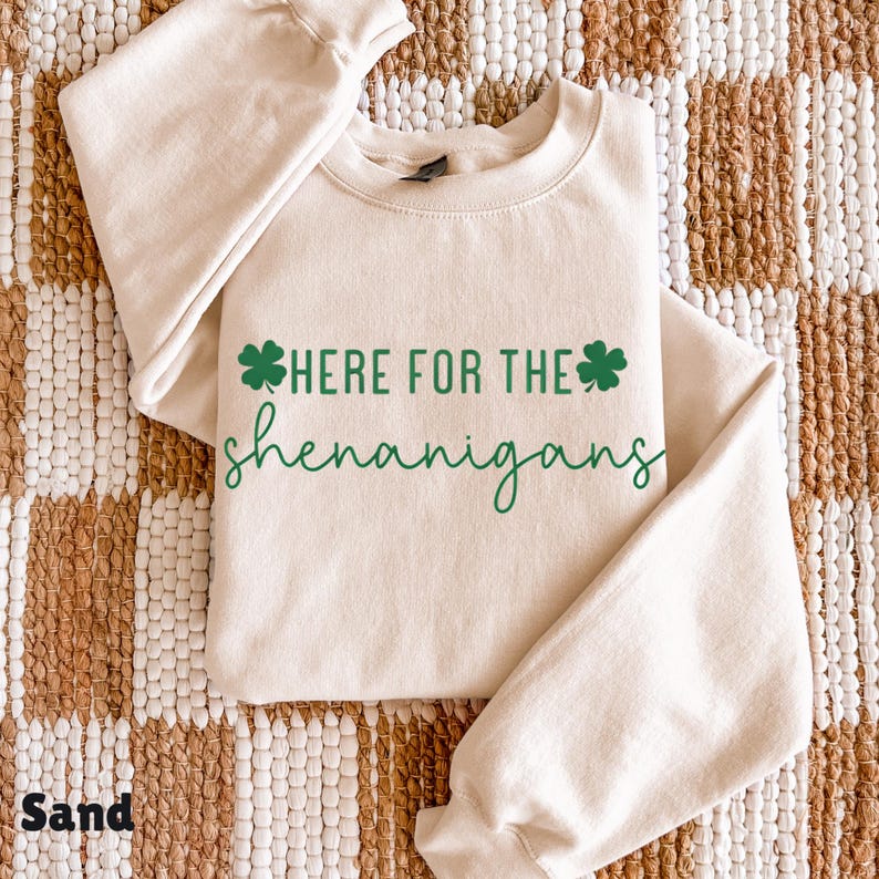 May include: A sand-colored sweatshirt with the text "HERE FOR THE shenanigans" in green, along with two green shamrock graphics. The sweatshirt is folded on a woven, tan and brown surface.