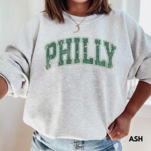 Op de afbeelding: Lichtgrijze sweatshirt met het woord "PHILLY" in versleten groene letters. De sweatshirt heeft een ronde hals en lange mouwen. Het woord "ASH" is rechtsonder in het zwart gedrukt.