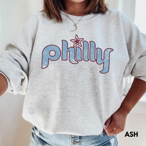 May include: Light gray sweatshirt with the word "Philly" in light blue, outlined in burgundy, and a star graphic. The sweatshirt is a crew neck style and has long sleeves. The word "ASH" is printed in black in the lower right corner.