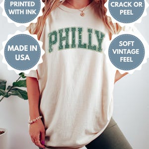 Op de afbeelding: Ivoor T-shirt met "PHILLY" gedrukt in verweerde groene letters. Het shirt heeft de tekst "DTG-PRINTED WITH INK", "MADE IN USA", "WON'T CRACK OR PEEL" en "SOFT VINTAGE FEEL".