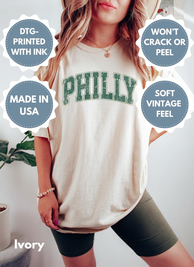 Op de afbeelding: Ivoor T-shirt met "PHILLY" gedrukt in verweerde groene letters. Het shirt heeft de tekst "DTG-PRINTED WITH INK", "MADE IN USA", "WON'T CRACK OR PEEL" en "SOFT VINTAGE FEEL".