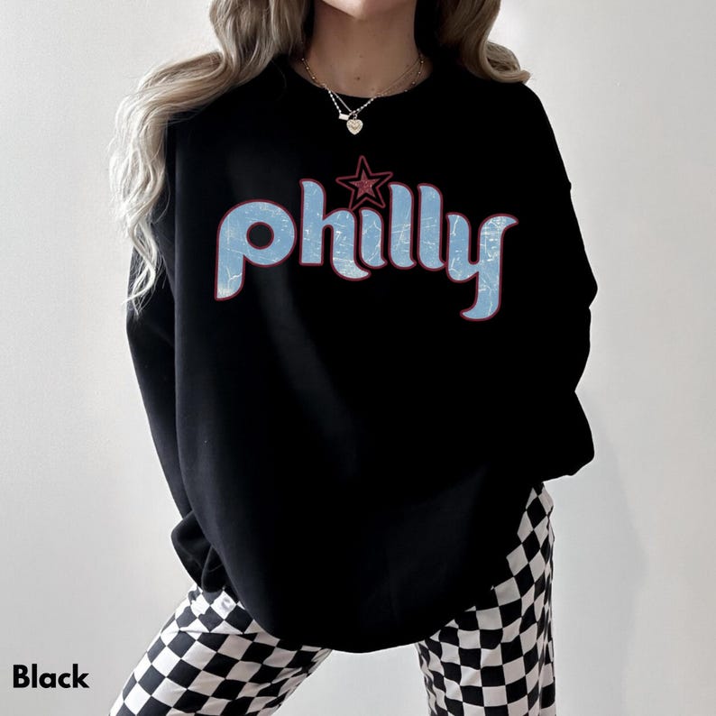 May include: Black crewneck sweatshirt with the word "philly" in light blue, outlined in red, and a red star above the word. The sweatshirt is worn with black and white checkered pants.