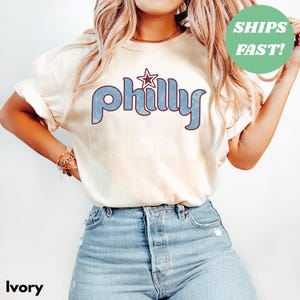May include: Ivory-colored t-shirt with the word "Philly" in blue, retro-style lettering and a star graphic. The shirt has rolled-up sleeves and is paired with blue jeans. A green circle with the text "SHIPS FAST!" is in the upper right corner.