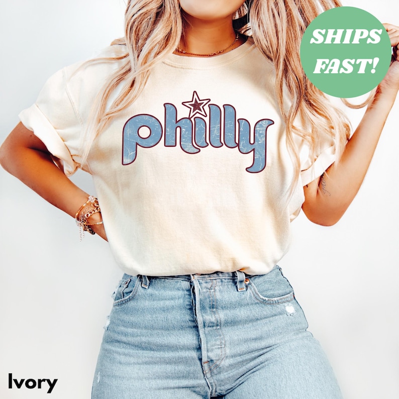 May include: Ivory-colored t-shirt with the word "Philly" in blue, retro-style lettering and a star graphic. The shirt has rolled-up sleeves and is paired with blue jeans. A green circle with the text "SHIPS FAST!" is in the upper right corner.