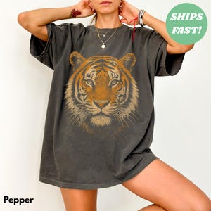 Peut inclure: T-shirt gris foncé avec un motif de visage de tigre détaillé en orange et marron. Le t-shirt a un aspect vintage et usé. Le mot "Pepper" est imprimé en bas à gauche.