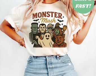 May include: Ivory-colored t-shirt featuring a graphic of classic Halloween monsters, including Frankenstein, Dracula, a mummy, a witch, a werewolf, and a ghost. The words "Monster Mash" are printed above the characters. The shirt is a casual, crew neck style.
