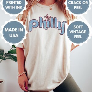 May include: Ivory-colored t-shirt with the word "Philly" in light blue, featuring a star graphic. The shirt has a soft, vintage feel and is made in the USA. The design is printed with ink that won't crack or peel.