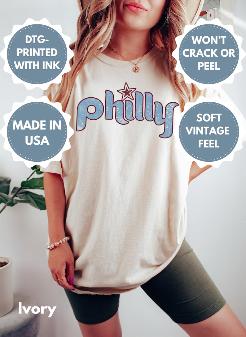May include: Ivory-colored t-shirt with the word "Philly" in light blue, featuring a star graphic. The shirt has a soft, vintage feel and is made in the USA. The design is printed with ink that won't crack or peel.