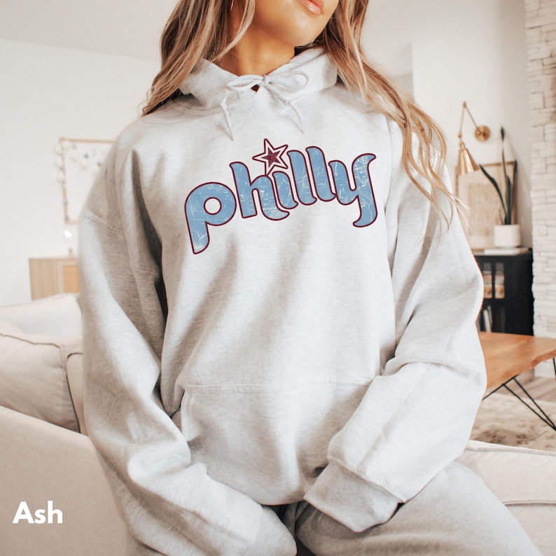 May include: Ash-colored hoodie with the word "Philly" in blue, outlined in burgundy, and a star graphic. The hoodie has a drawstring hood and a front pocket. The word "Ash" is printed at the bottom left.