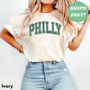 Op de afbeelding: Ivoor kleurig t-shirt met het woord "PHILLY" in verweerde groene letters. Het shirt wordt gecombineerd met lichtblauwe denim jeans. Een groene cirkel met de tekst "SHIPS FAST!" is ook zichtbaar.
