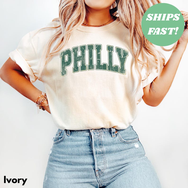 Op de afbeelding: Ivoor kleurig t-shirt met het woord "PHILLY" in verweerde groene letters. Het shirt wordt gecombineerd met lichtblauwe denim jeans. Een groene cirkel met de tekst "SHIPS FAST!" is ook zichtbaar.