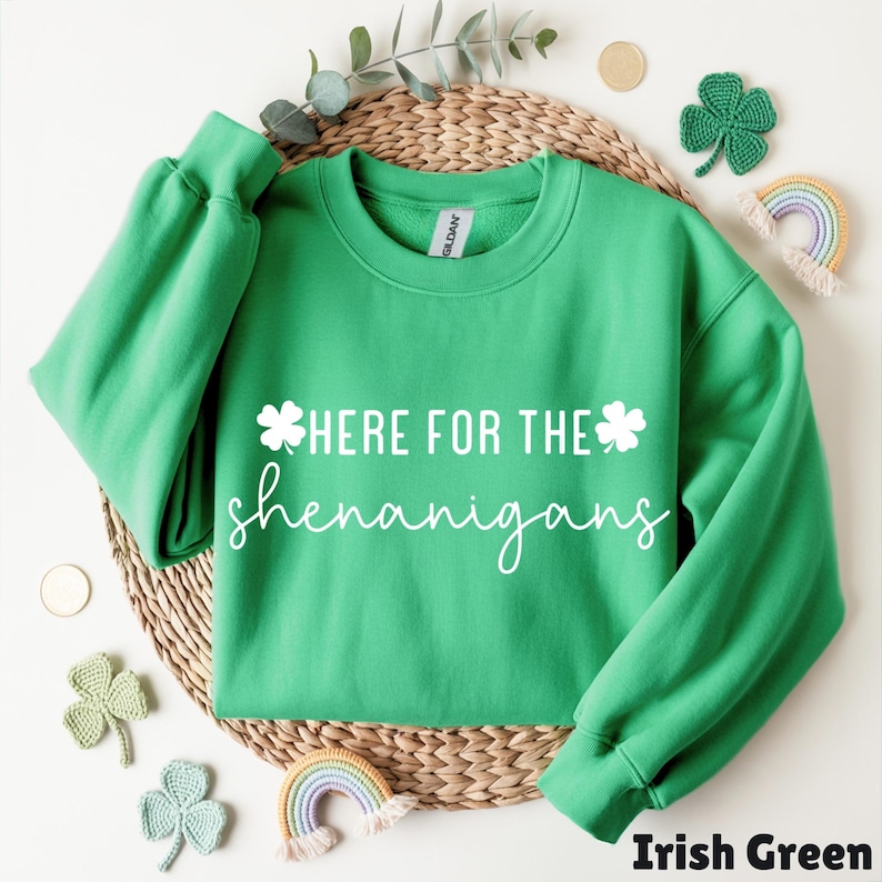 May include: Irish green sweatshirt with the text "HERE FOR THE shenanigans" in white script. The sweatshirt is displayed on a woven mat with decorative shamrocks and a rainbow.