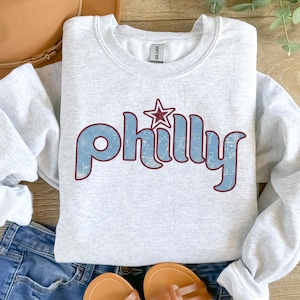 May include: Ash gray crewneck sweatshirt with the word "Philly" in light blue, outlined in burgundy, and a burgundy star. The sweatshirt is folded on top of blue denim shorts, with a brown hat and sandals.