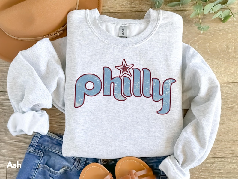 May include: Ash gray crewneck sweatshirt with the word "Philly" in light blue, outlined in burgundy, and a burgundy star. The sweatshirt is folded on top of blue denim shorts, with a brown hat and sandals.