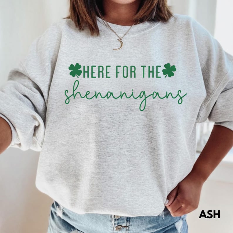 May include: A light gray sweatshirt with the text "HERE FOR THE shenanigans" in green, with shamrock graphics. The word "ASH" is printed in the bottom right corner.