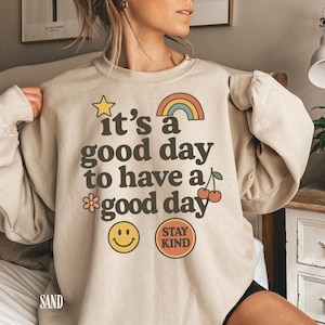 May include: Sand-colored sweatshirt with the phrase "it's a good day to have a good day" in black lettering. The design includes a yellow star, rainbow, flower, smiley face, cherries, and a "Stay Kind" badge.