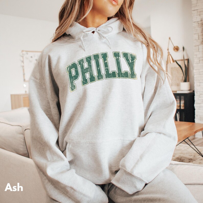 Op de afbeelding: Een lichtgrijze hoodie met het woord "PHILLY" in verweerde groene letters over de borst. De hoodie heeft een capuchon met trekkoord en een kangoeroezak. Het woord "Ash" staat linksonder in het wit gedrukt.