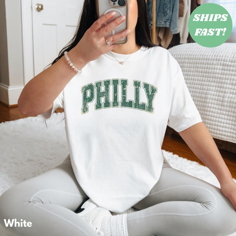 Op de afbeelding: Wit T-shirt met het woord "PHILLY" in verweerde groene letters. Het shirt heeft een ronde hals en korte mouwen. De tekst is in een lettertype in collegestijl. Het shirt is gemaakt van een zacht, comfortabel materiaal.