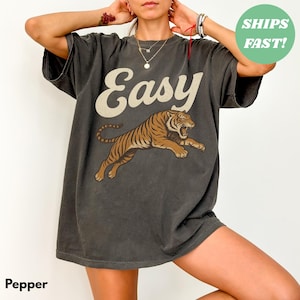 May include: Dark grey t-shirt with the word "Easy" in cream-colored script above a leaping tiger illustration in brown and black. The shirt is a relaxed fit.