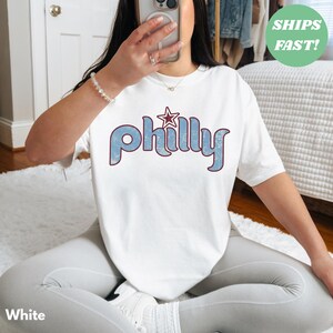 May include: White t-shirt with the word "Philly" in blue, outlined in maroon, and a star graphic. The shirt is a casual style.