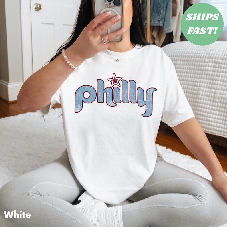 May include: White t-shirt with the word "Philly" in blue, outlined in maroon, and a star graphic. The shirt is a casual style.