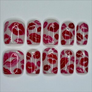 Puede incluir: Un conjunto de diez u&ntilde;as artificiales con una base transparente, adornadas con un patr&oacute;n repetido de marcas de besos de l&aacute;piz labial rojo. Las u&ntilde;as est&aacute;n dispuestas en dos filas, mostrando las vibrantes huellas de labios.