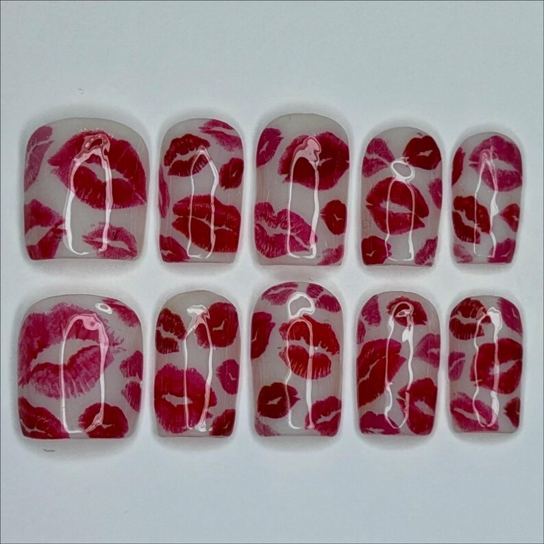 Puede incluir: Un conjunto de diez u&ntilde;as artificiales con una base transparente, adornadas con un patr&oacute;n repetido de marcas de besos de l&aacute;piz labial rojo. Las u&ntilde;as est&aacute;n dispuestas en dos filas, mostrando las vibrantes huellas de labios.