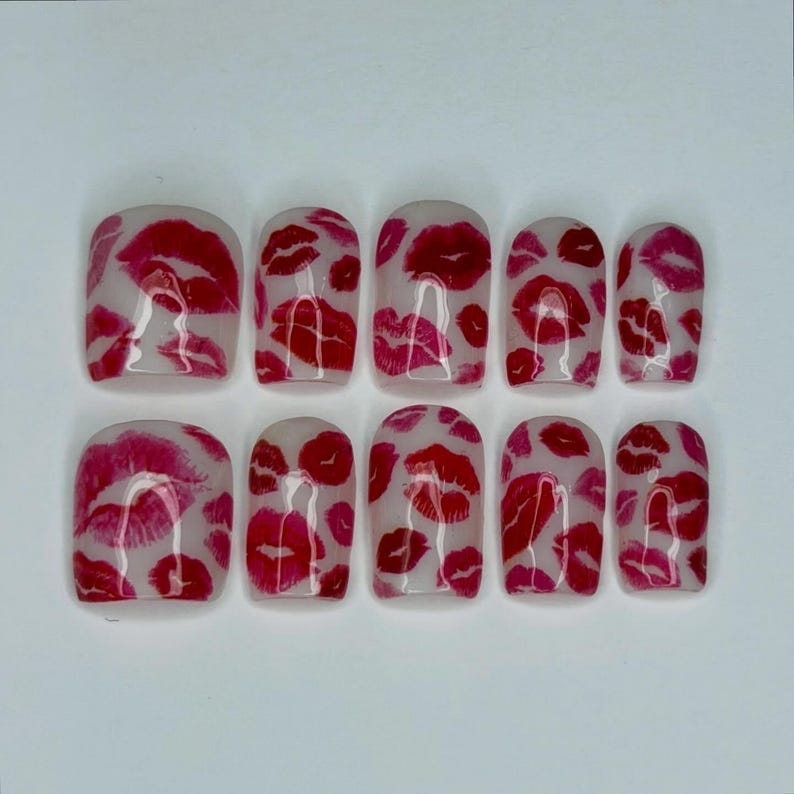 Puede incluir: Un conjunto de diez u&ntilde;as artificiales con una base transparente y un patr&oacute;n repetido de marcas de besos de l&aacute;piz labial rojo. Las u&ntilde;as son de forma cuadrada y est&aacute;n dispuestas en dos filas de cinco.