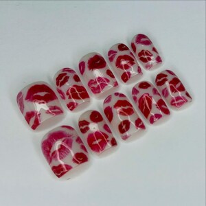 Puede incluir: Un conjunto de diez u&ntilde;as postizas con una base transparente y un patr&oacute;n repetido de labios rojos y rosas. Las u&ntilde;as son de forma rectangular y est&aacute;n dispuestas sobre una superficie blanca.