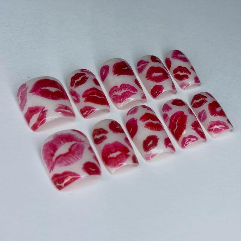 Puede incluir: Un conjunto de diez u&ntilde;as artificiales con una base blanca y un patr&oacute;n repetido de marcas de besos de l&aacute;piz labial rojo y rosa. Las u&ntilde;as tienen varias formas, incluyendo cuadradas y ovaladas.