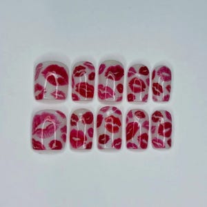 Puede incluir: Un conjunto de diez u&ntilde;as postizas con base transparente, cada una adornada con un patr&oacute;n de huellas de labios rojos y rosas. Las u&ntilde;as est&aacute;n dispuestas en dos filas de cinco, mostrando un dise&ntilde;o audaz y juguet&oacute;n.