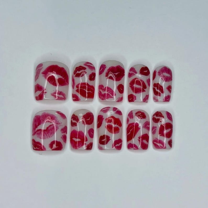 Puede incluir: Un conjunto de diez u&ntilde;as postizas con base transparente, cada una adornada con un patr&oacute;n de huellas de labios rojos y rosas. Las u&ntilde;as est&aacute;n dispuestas en dos filas de cinco, mostrando un dise&ntilde;o audaz y juguet&oacute;n.