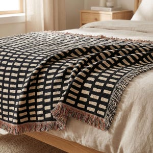 May include: A black and beige woven throw blanket with a geometric pattern of rectangular blocks. The blanket is draped over a bed, showcasing its fringed edges and textured design. The color palette is neutral, with a touch of red at the fringe.