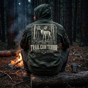 May include: A camouflage print hooded sweatshirt featuring a graphic on the back of a skeletal deer in a forest. The text reads TRAIL CAM TERROR, WHATEVER IT WAS, IT HAD ANTLERS, 41°F, 10/28/24, and 02:13 AM, with a small deer icon in the corner.
