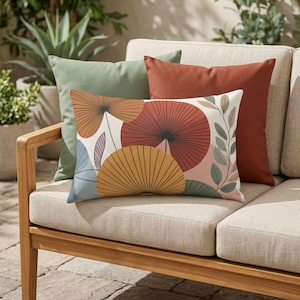 May include: A set of decorative throw pillows in shades of green, rust, and beige. One pillow features a botanical design with orange, yellow, and blue fan-like shapes. The pillows are arranged on a light-colored outdoor sofa.