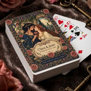 May include: A deck of playing cards featuring an ornate illustration of two figures with long hair, surrounded by roses and foliage. The text reads Clara & June, IN BLOOM SINCE 2008, and BROOKLYN, NEW YORK. Several playing cards are fanned out behind the deck.