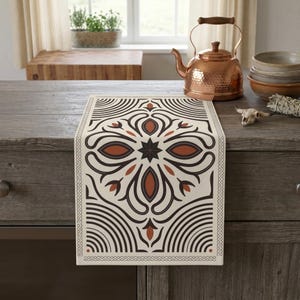 May include: A rectangular table runner featuring a cream-colored background with a symmetrical, geometric pattern in dark brown and burnt orange. The design includes central star and petal motifs surrounded by curved lines and a border.