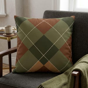 May include: A square throw pillow featuring an argyle pattern in shades of olive green, dark forest green, and terracotta brown. The design includes thin, dashed white lines forming a diamond grid across the surface of the pillow.