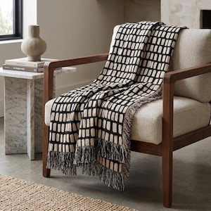 May include: A woven throw blanket featuring a repeating pattern of black rectangular blocks on a cream background, finished with fringed edges. The blanket is draped over a wooden armchair with light-colored upholstery and a matching cream cushion.
