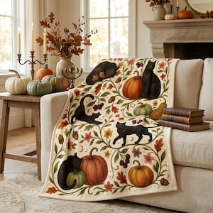 Könnte beinhalten: Eine cremefarbene Wohndecke mit einem wiederkehrenden Muster aus schwarzen Katzen, orangefarbenen Kürbissen, grünen Zierkürbissen, Eicheln, Beeren und Herbstblättern. Das Design zeigt geschwungene Ranken und kleine Mondsicheln auf dem Stoff.