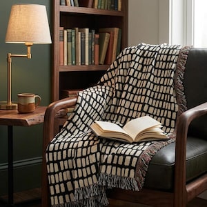 May include: A woven throw blanket featuring a repeating black and cream grid pattern with fringed edges. The blanket is draped over a dark leather armchair and has an open book resting on its surface, showcasing the textured fabric construction.
