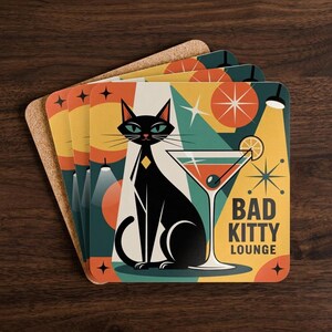 May include: A set of square cork-backed coasters featuring a graphic illustration of a black cat sitting next to a martini glass. The design includes geometric shapes, starbursts, and the text "BAD KITTY LOUNGE" in a bold, dark sans-serif font.