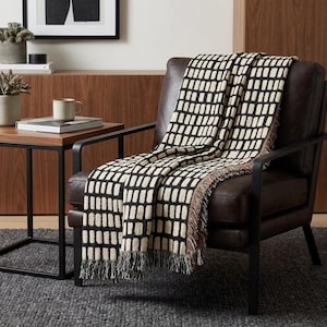 May include: A woven throw blanket featuring a repeating pattern of vertical cream-colored rectangular blocks against a solid black background. The blanket has a fringed edge along the bottom and is draped over a dark brown leather armchair.